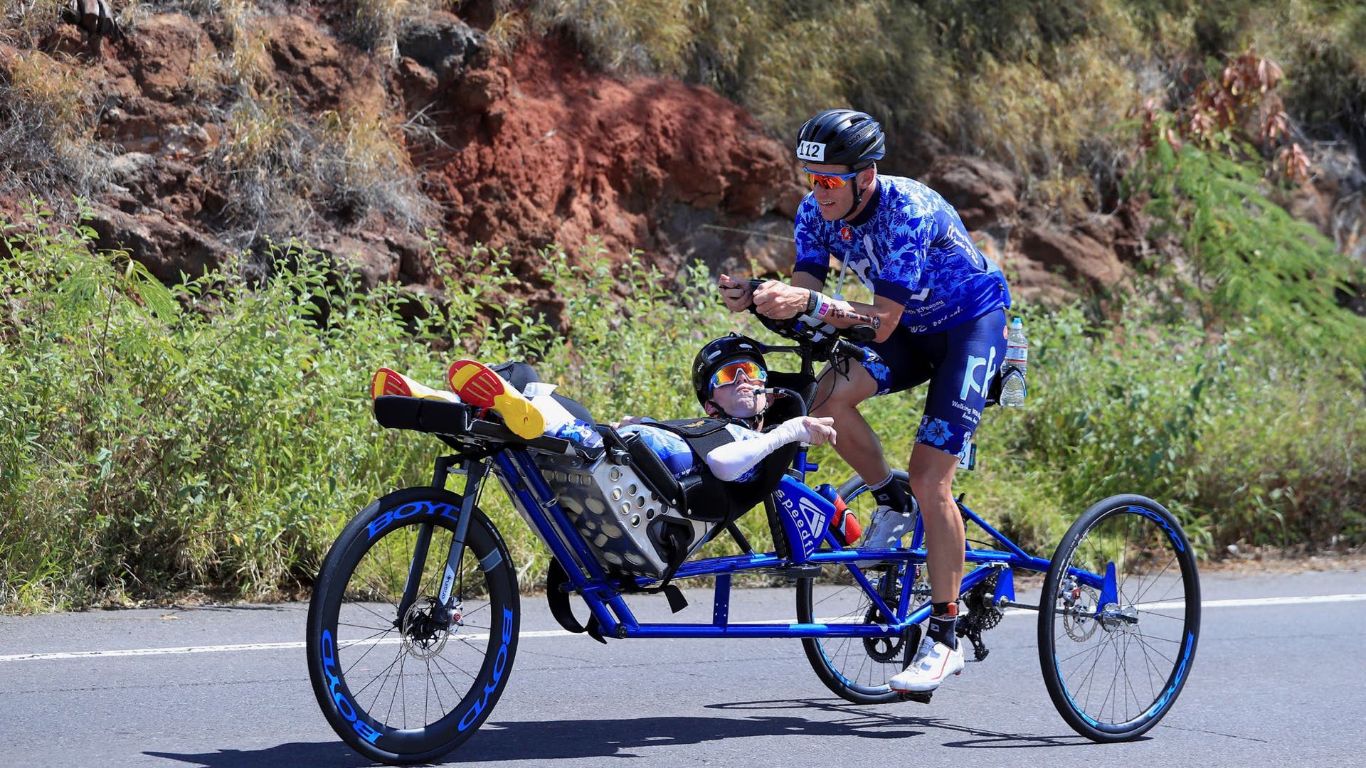A male triathlete pedals a tandem bike with a seat on the front, where another male triathlete is reclining.