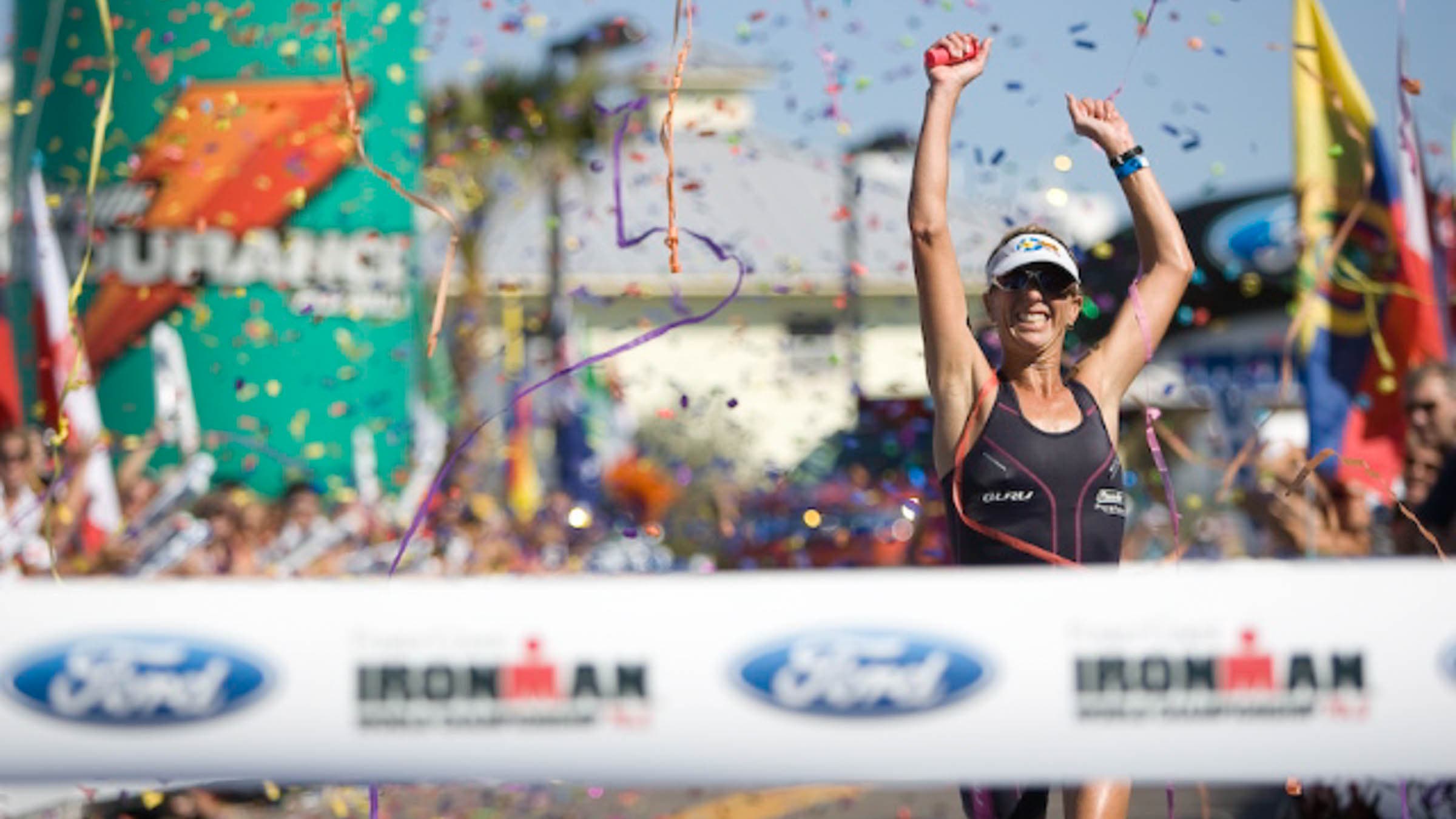 A woman runs triumphantly to the Ironman finish tape, arms raised