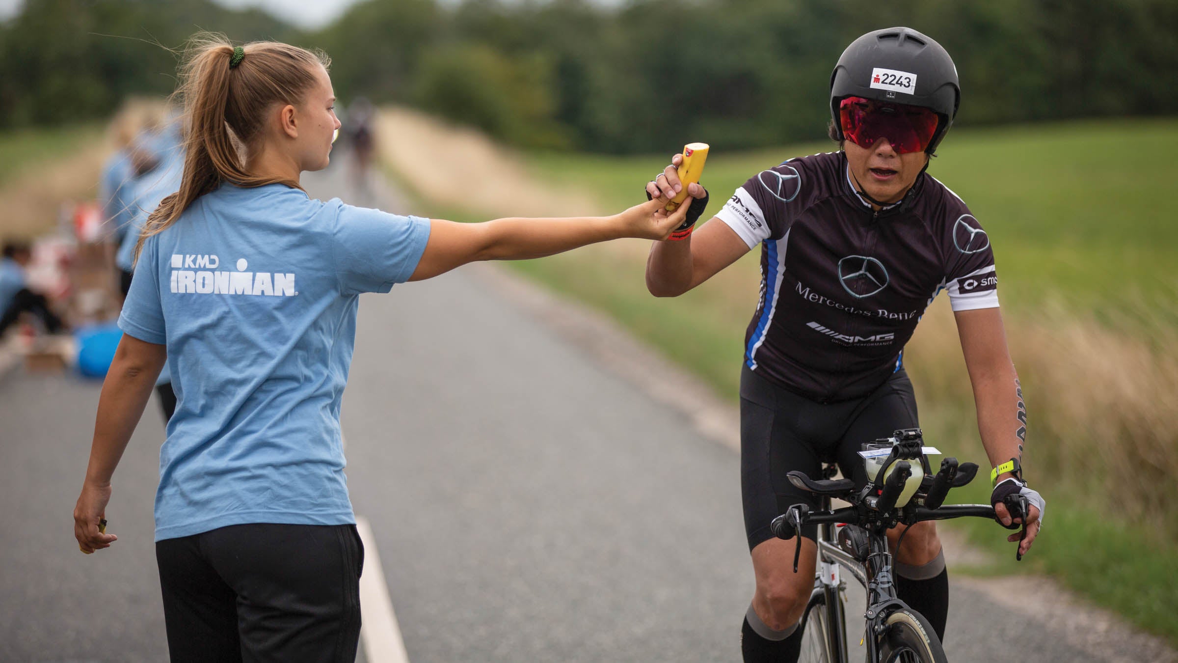 An athlete competes as a volunteer gives her a banana at an aid station