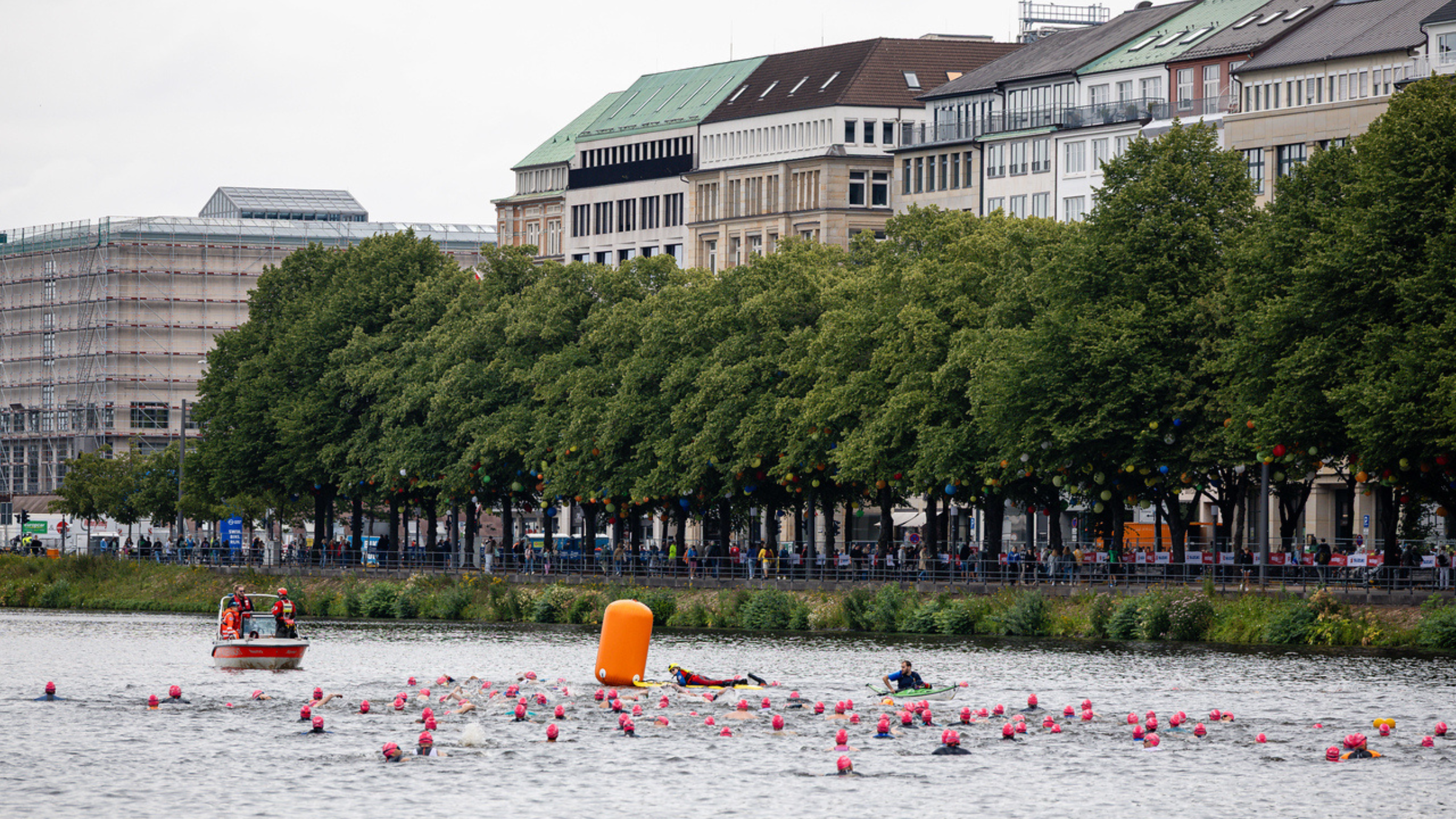athletes swim in wetsuits around an orange buoy in the middle of an urban area in Hamburg with buildings and trees along the water's edge