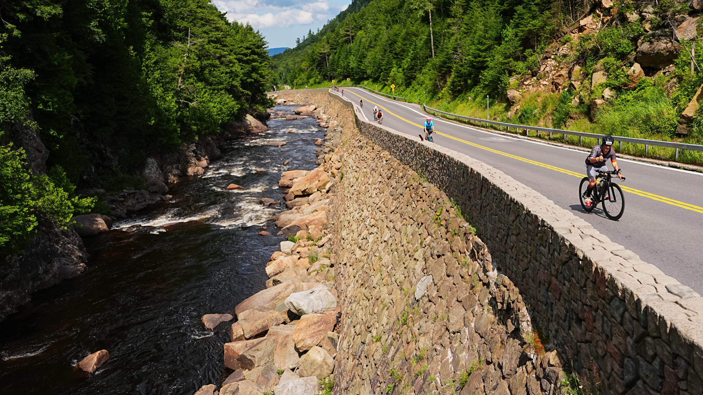 cyclists ride along wall overlooking river at Ironman Lake Placid