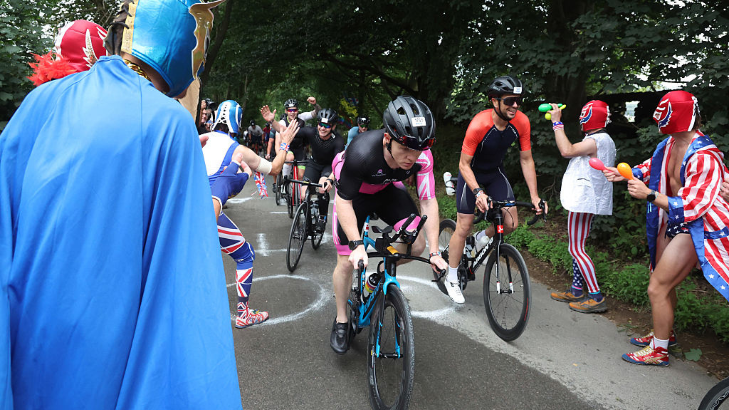 Ironman triathletes on bikes ride uphill on pavement surrounded by spectators in costumes cheering them on