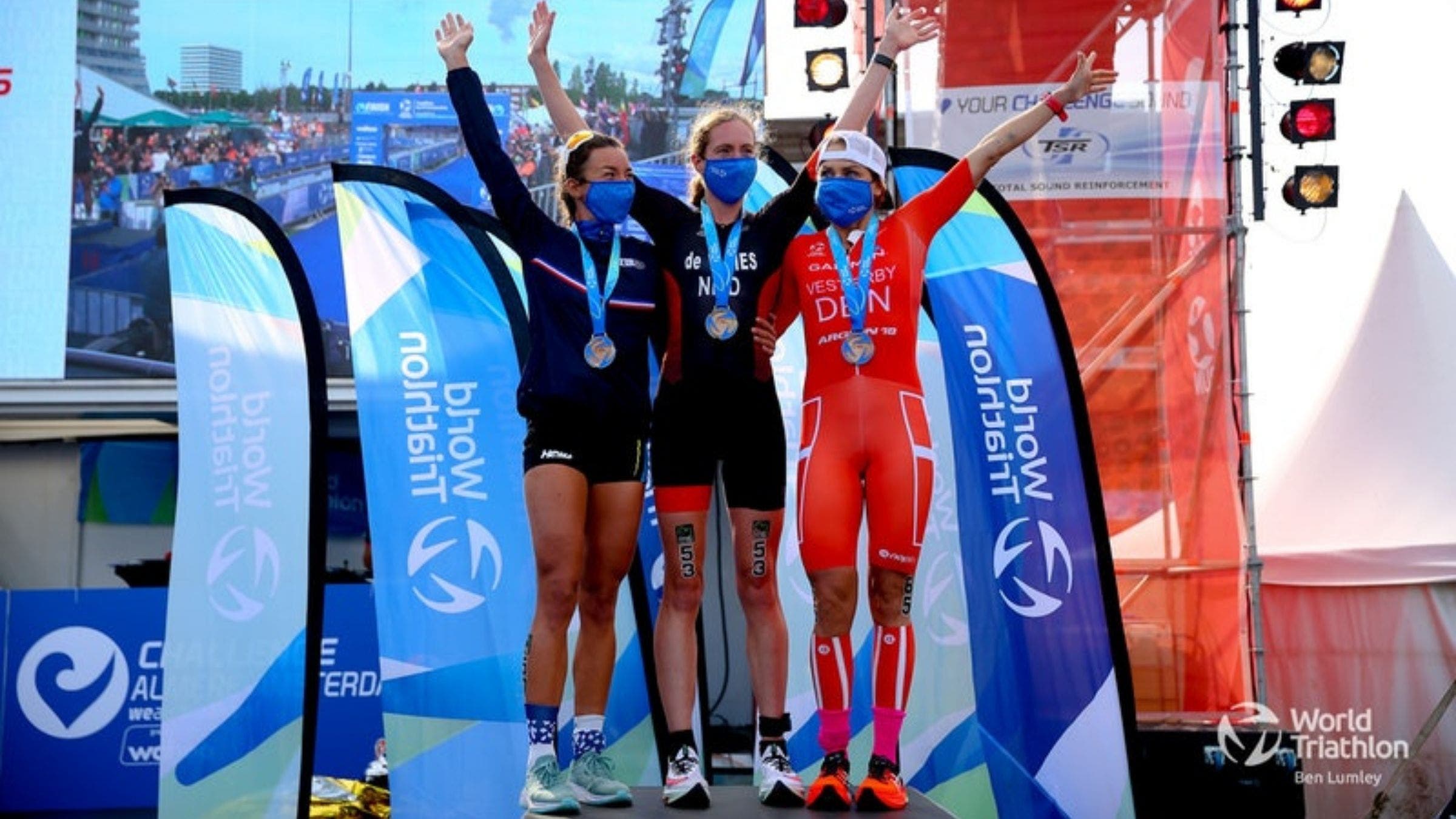 Three women stand atop the steps of a podium. They are wearing triathlon kits.