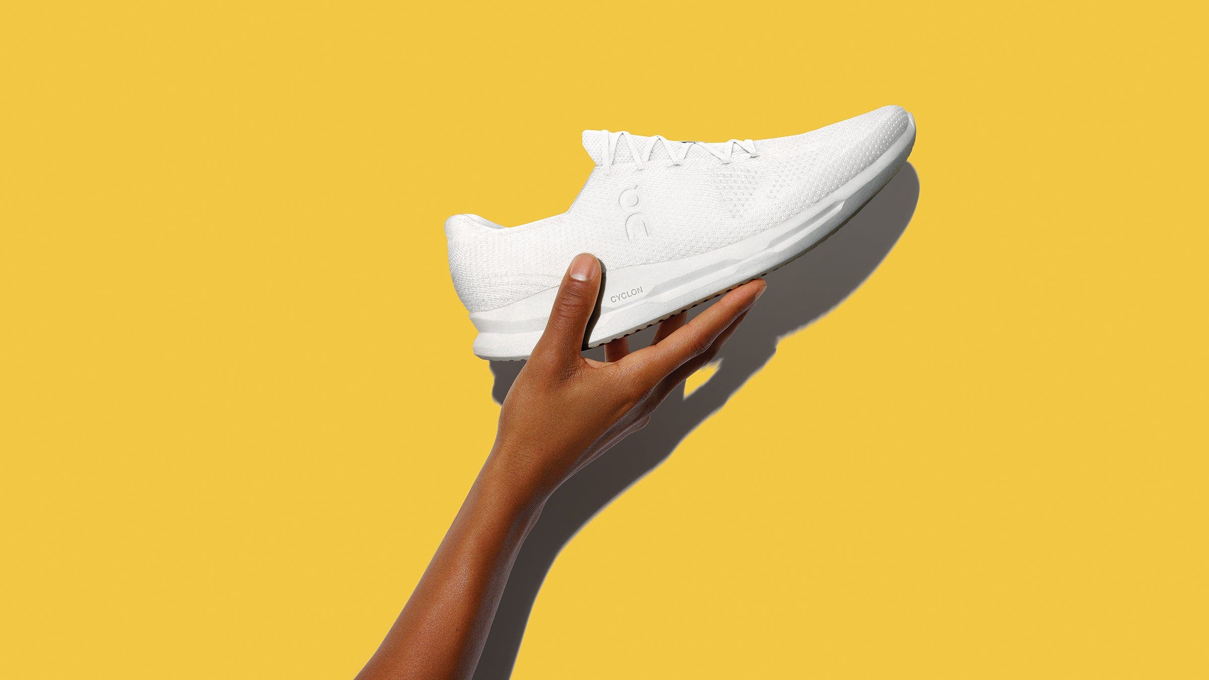 Hand holding white On Cyclon shoes, a sustainable running shoe, on a yellow background