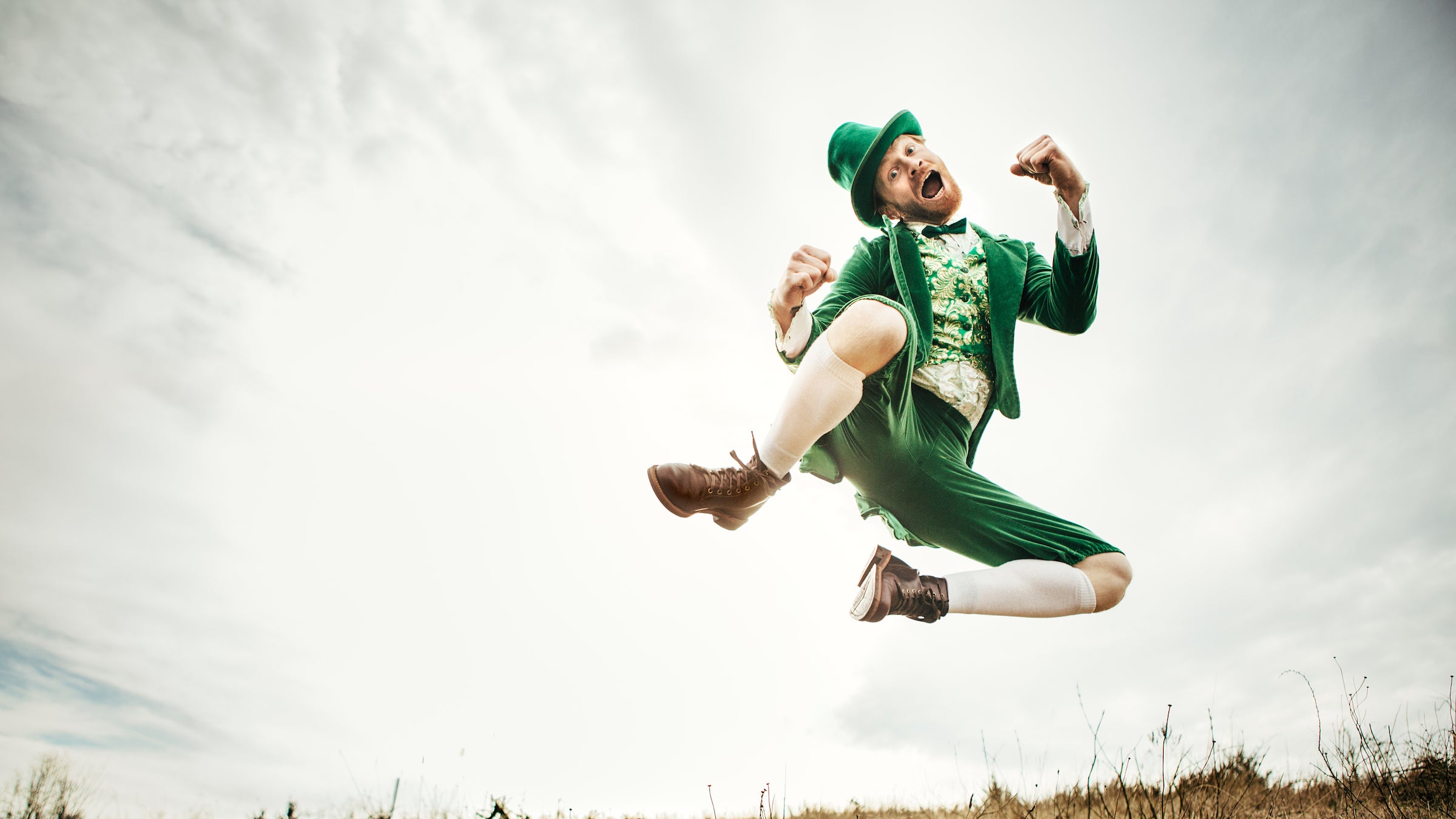 A stereotypical Irish character all ready for Saint Patricks day jumps and dances in an open field of Irish country side.  Copy space in the sky and grass.