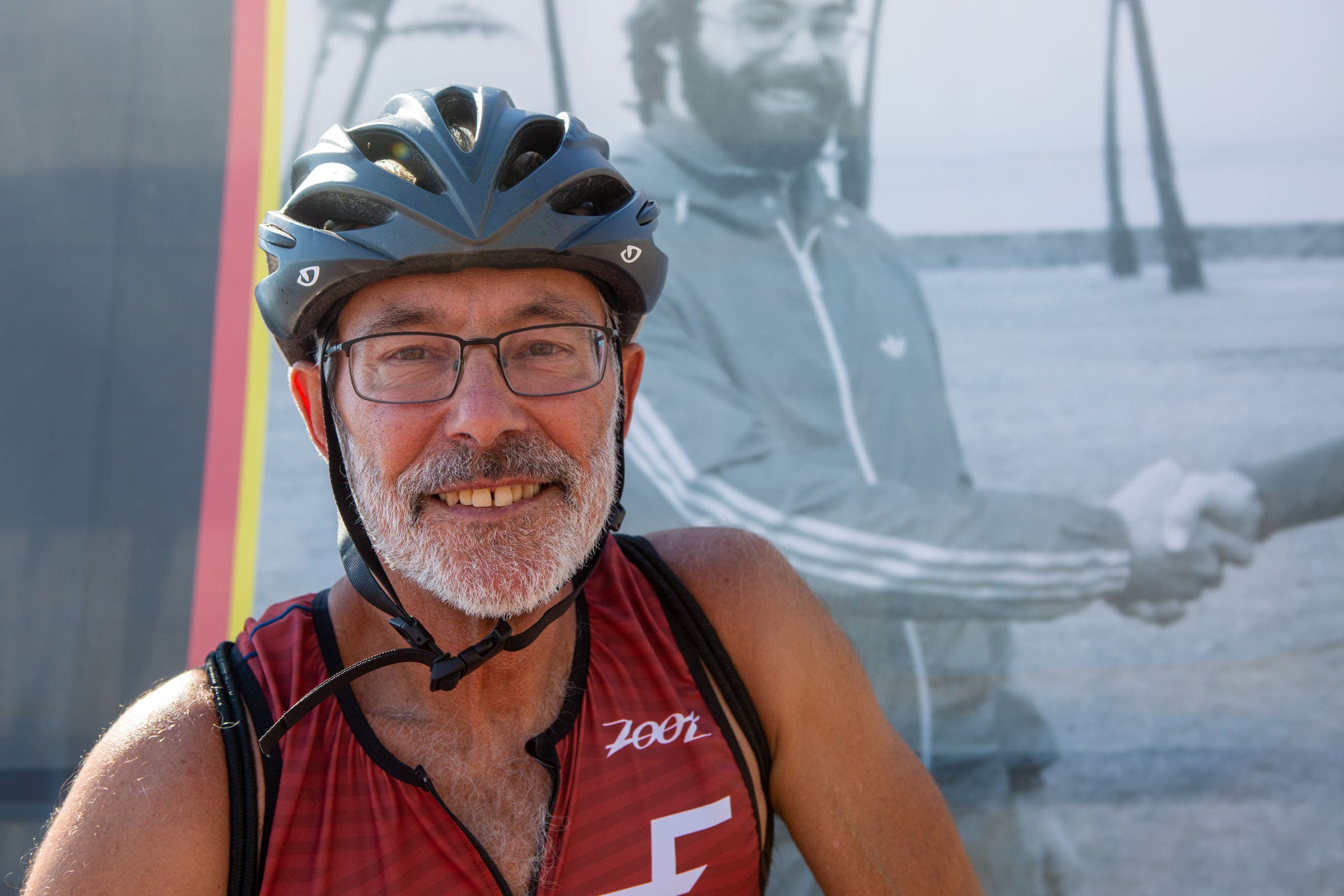 Gordon Haller, the first Ironman champion, smiles in front of a photo of himself at the 1987 Ironman World Championship