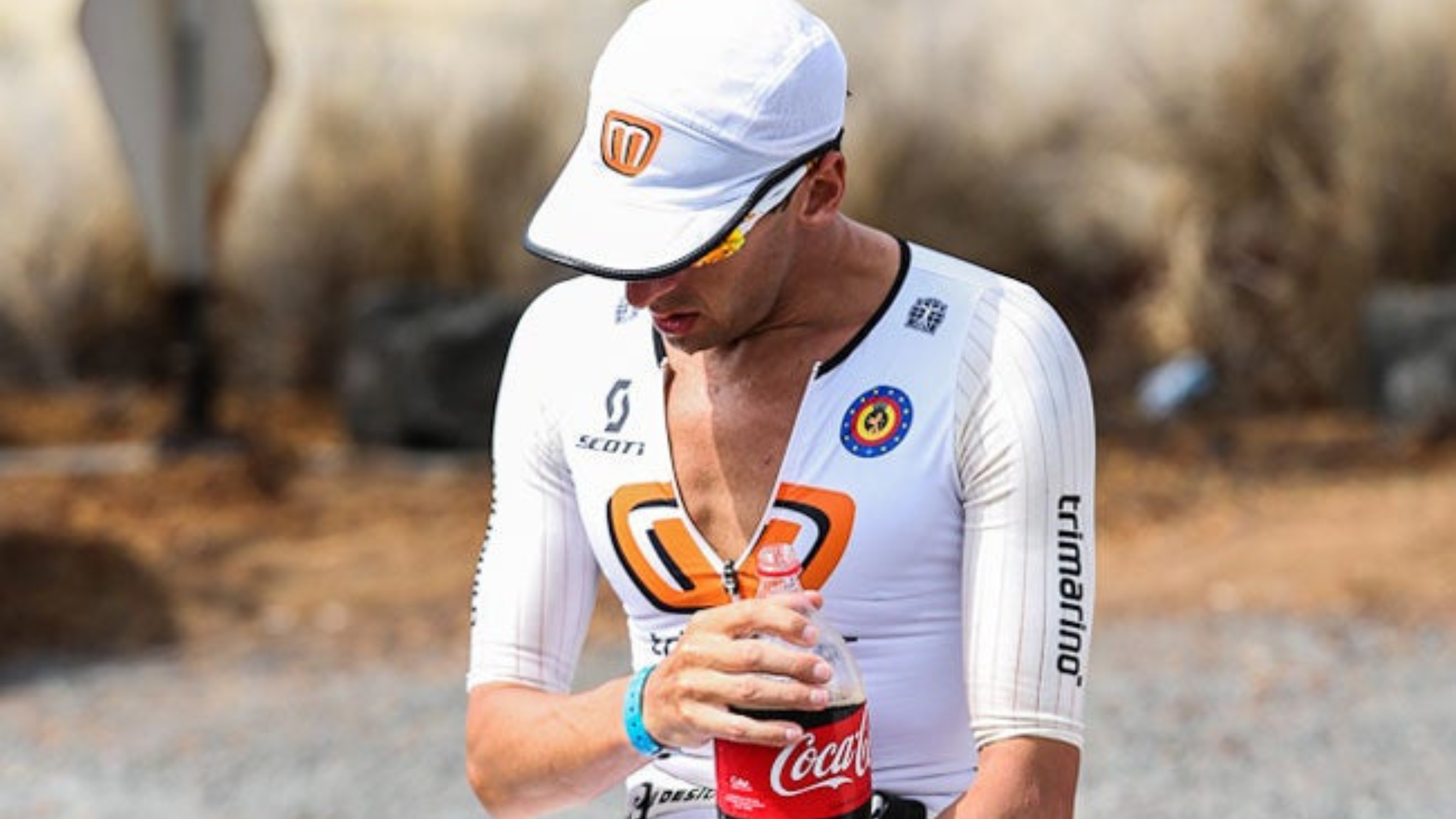 Man in white tri kit and hat sits down on a table with large bottle of Coca-Cola