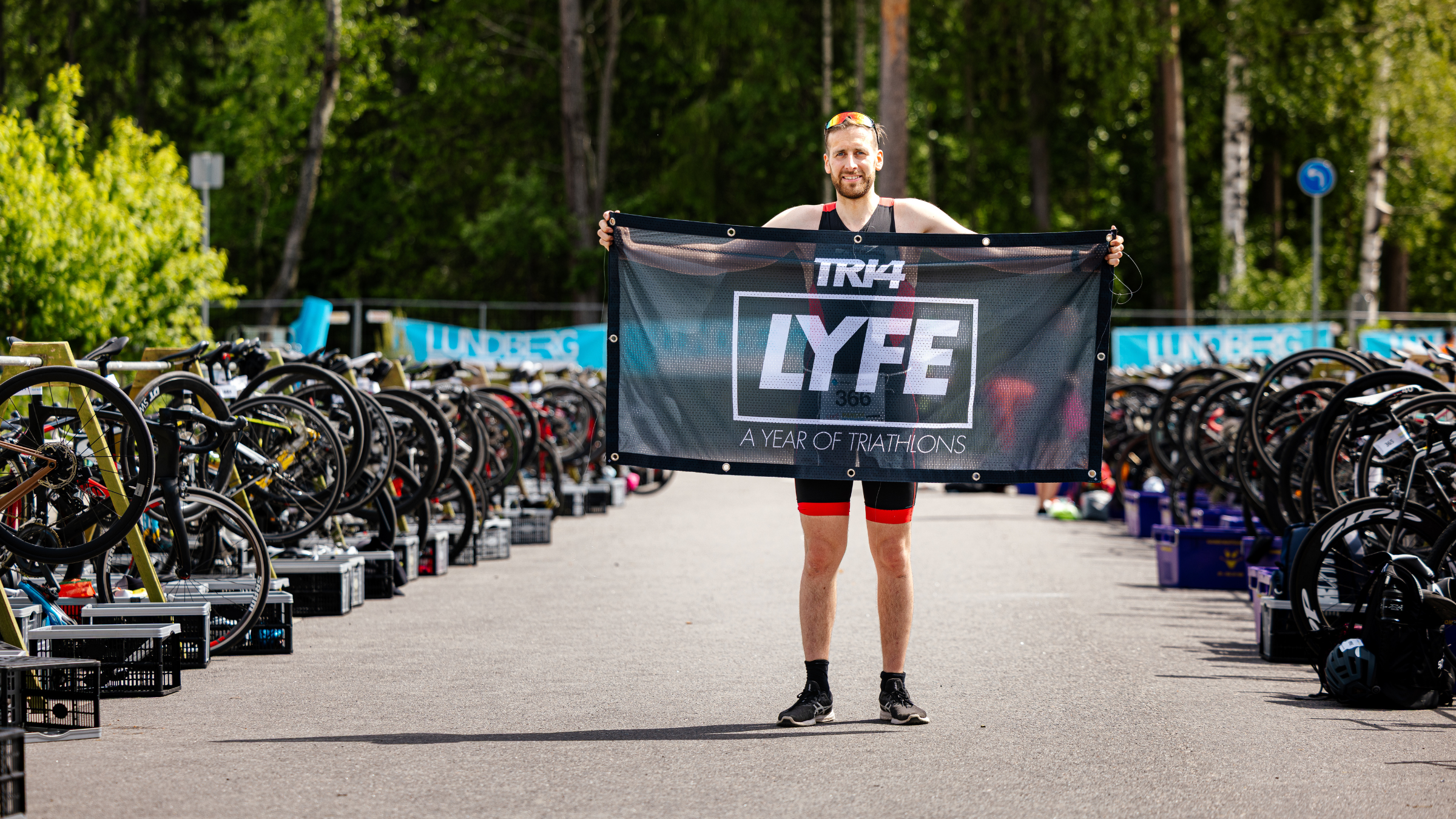 man stands in tri kit in transition area holding "Tri4Lyfe" banner with goggles on head