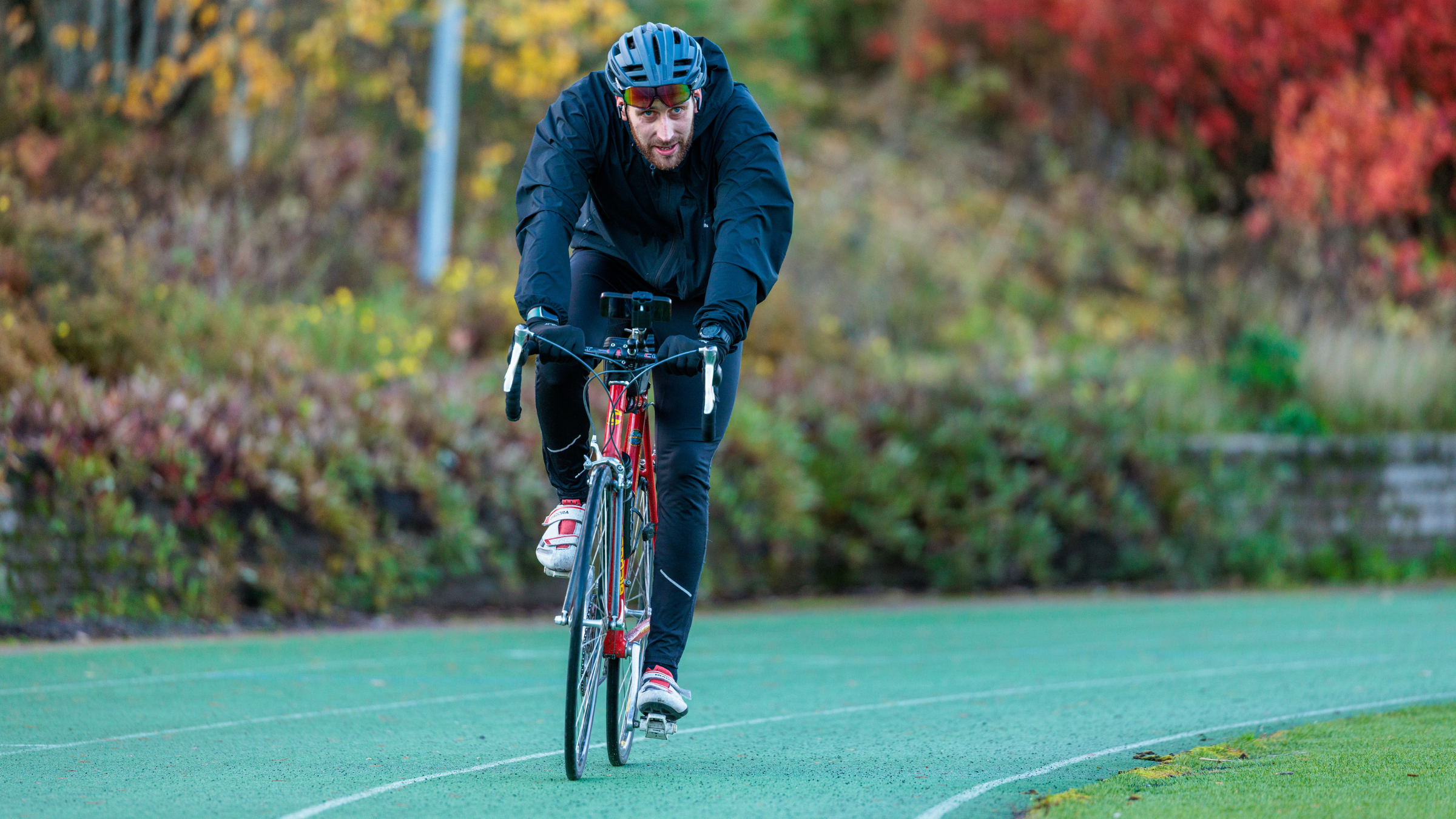 a man rides a road bike toward the camera dressed in winter gear and helmet on green road surface