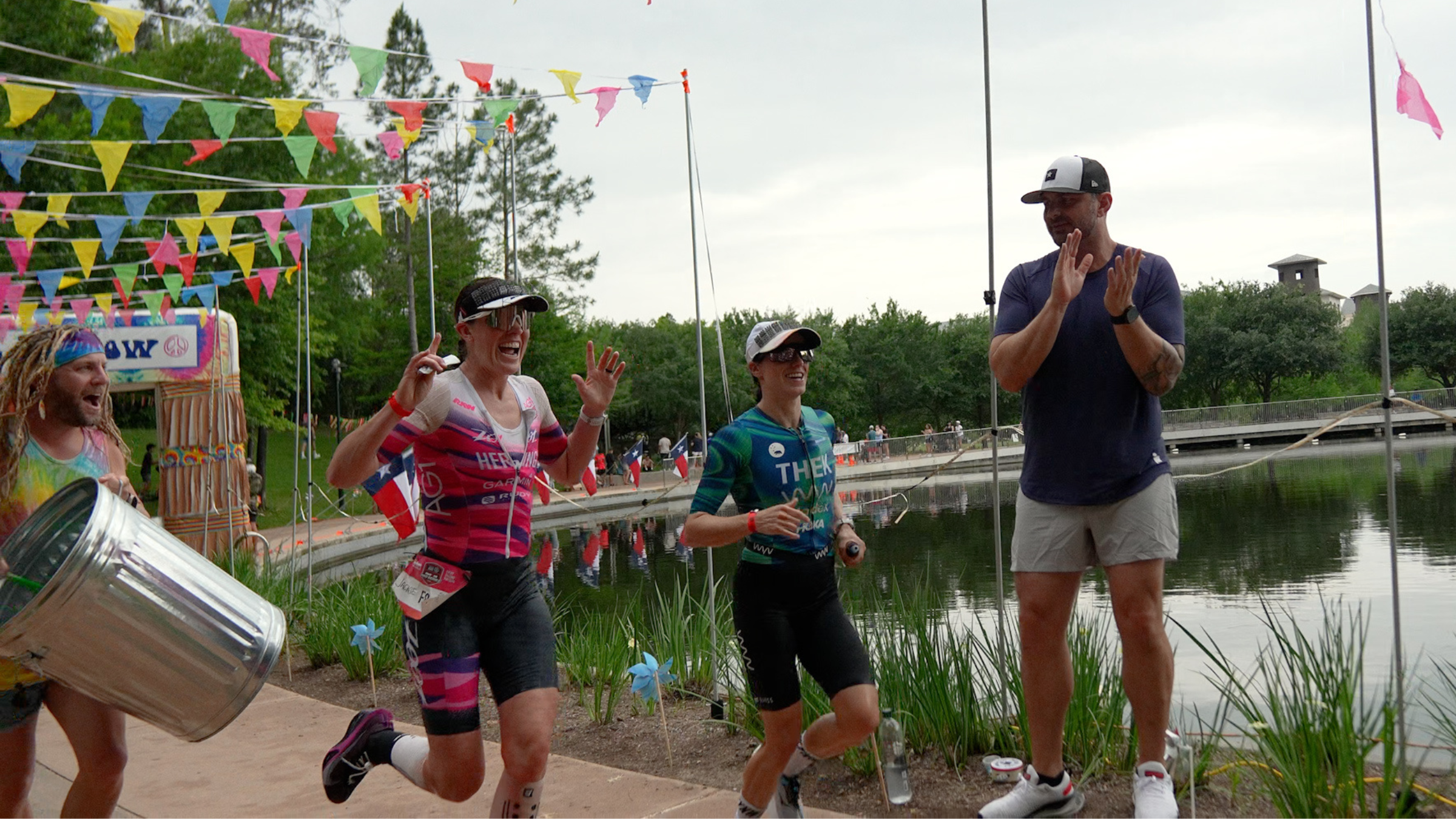 Two professional women triathletes run along the waterfront with cheering spectators, colorful flag banners raised above their heads.