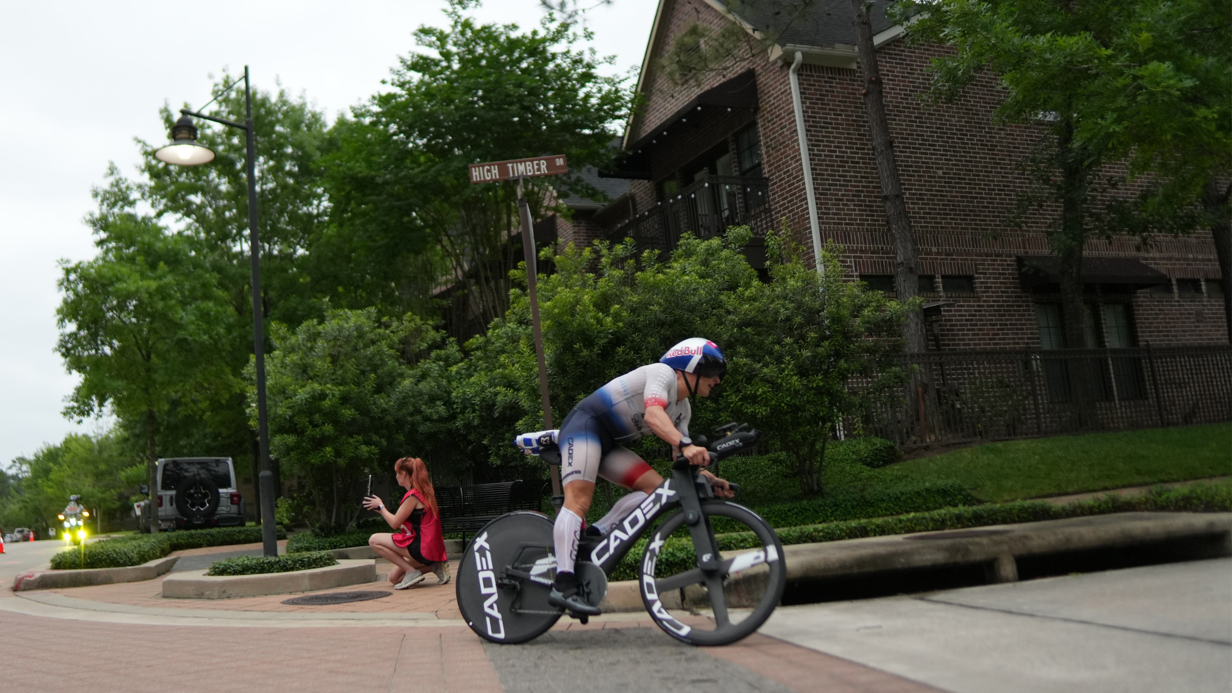 Man wearing professional triathlon kit riding triathlon bike with disc wheel around a corner with trees and houses in the background