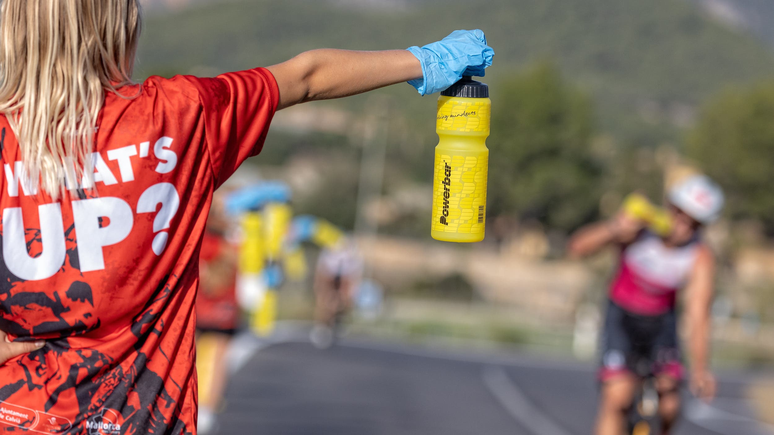 An aid station volunteer offers a bottle of sports drink to a triathlete on a bike.