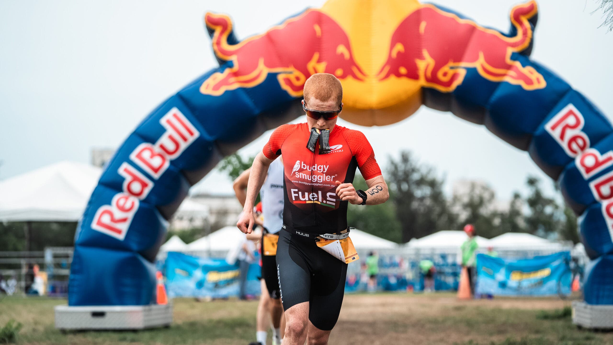 An athlete runs under a Red Bull inflatable arch during a race with two gel packs hanging from his mouth.