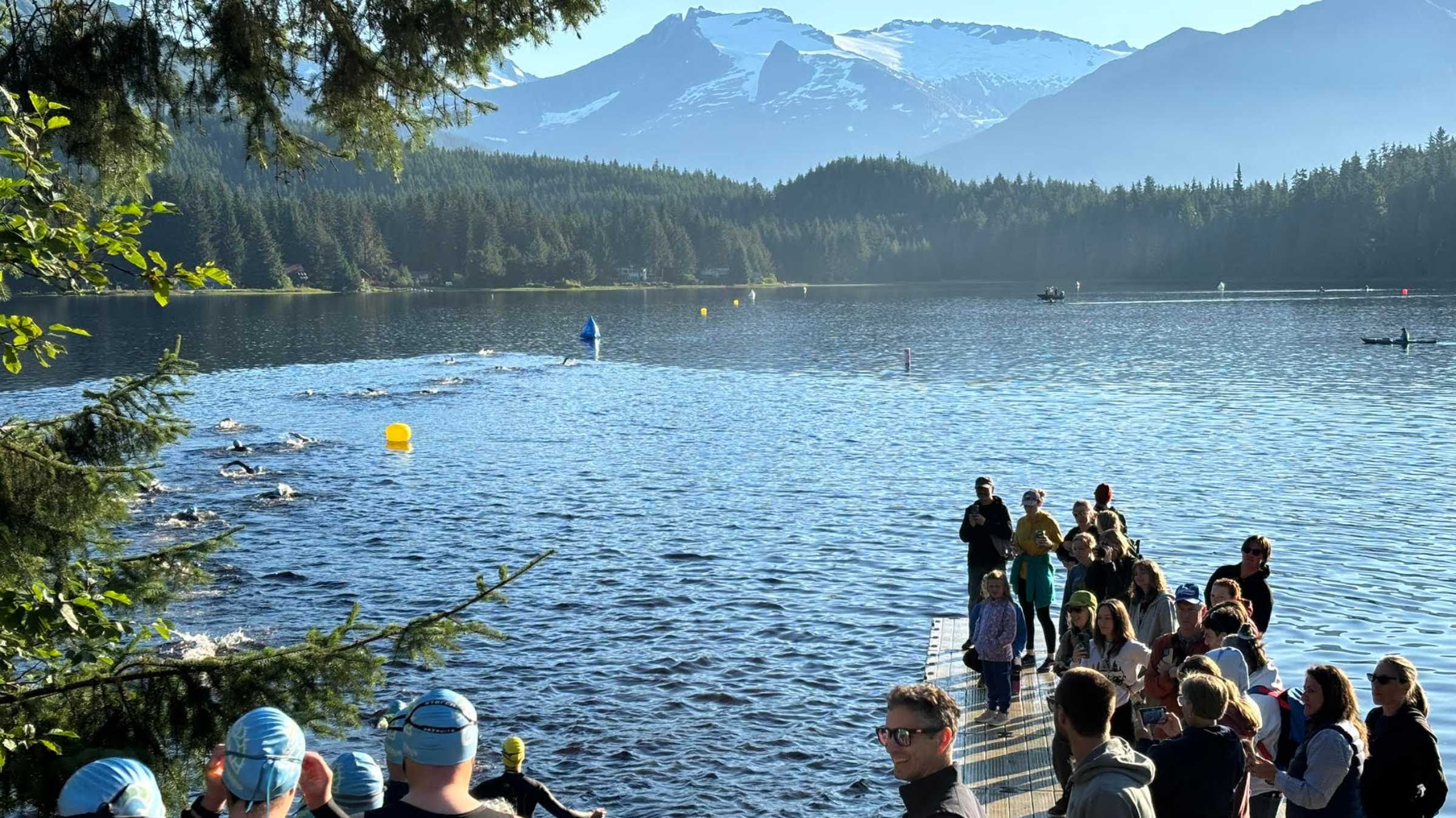 Triathletes in wetsuits and blue swim caps walk towards the lake with forests and mountains surrounding the lake and spectators on the pier, Aukeman Triathlon