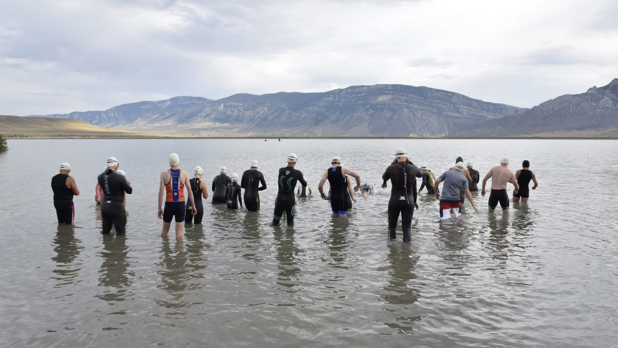 Triathletes in wetsuits wade into the water at the start of the Yellowstone Triathlon gates with mountains in the background.