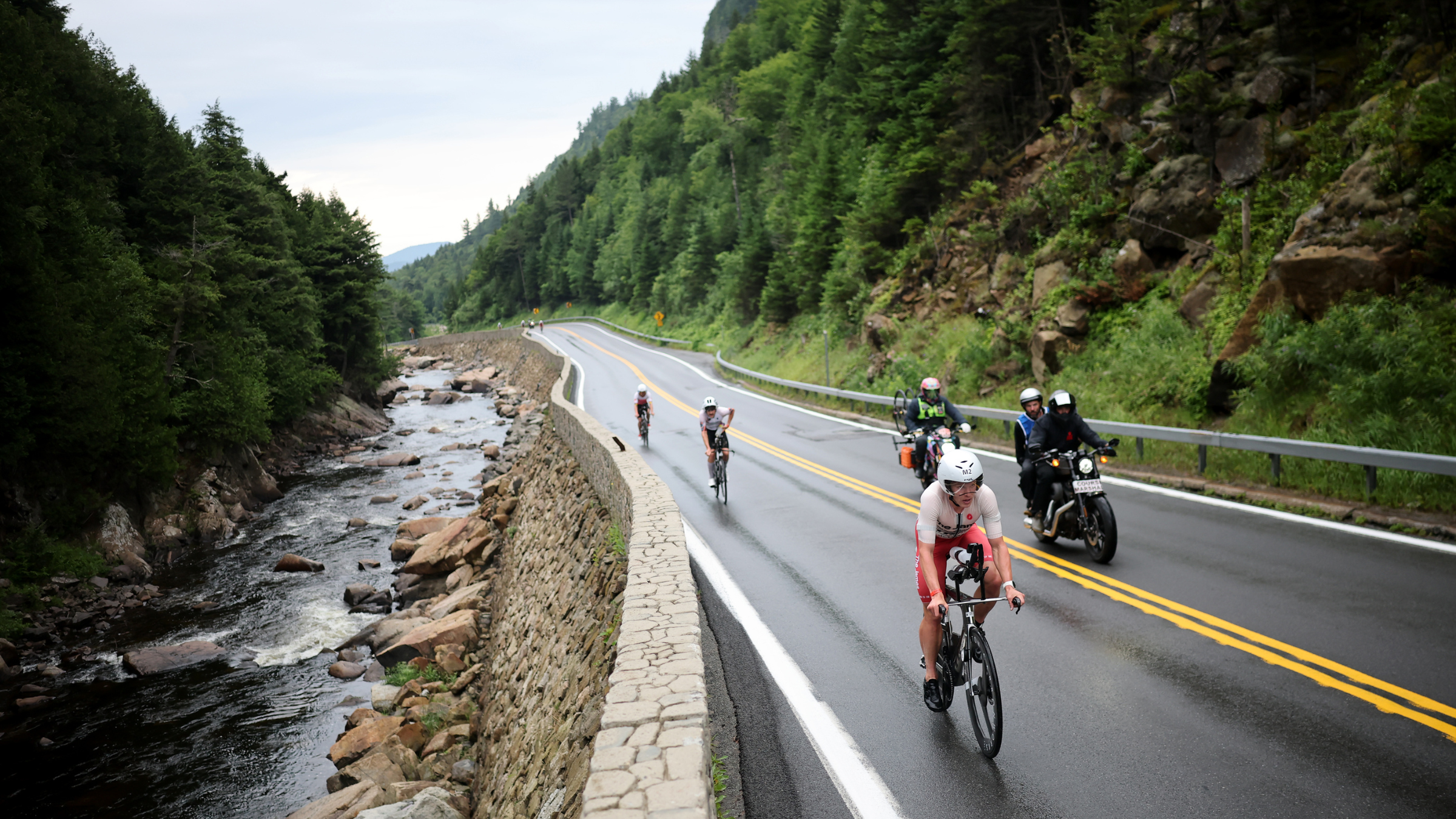 Cyclists ride triathlon bikes on the road through the valley with the river on one side and steep green hills on the other