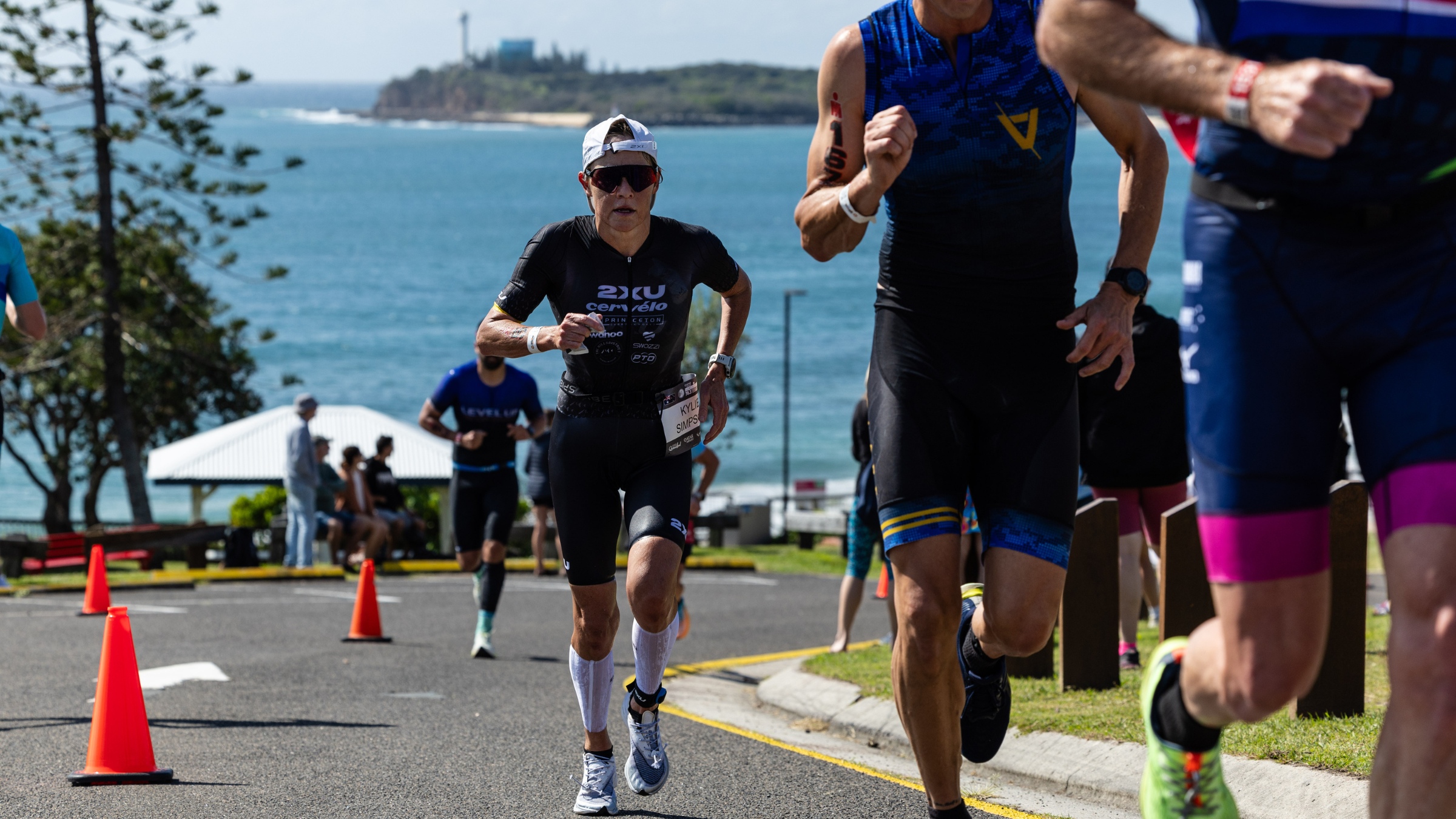 Athletes run uphill toward the camera on a route with the island and water in the background at Ironman 70.3 Sunshine Coast