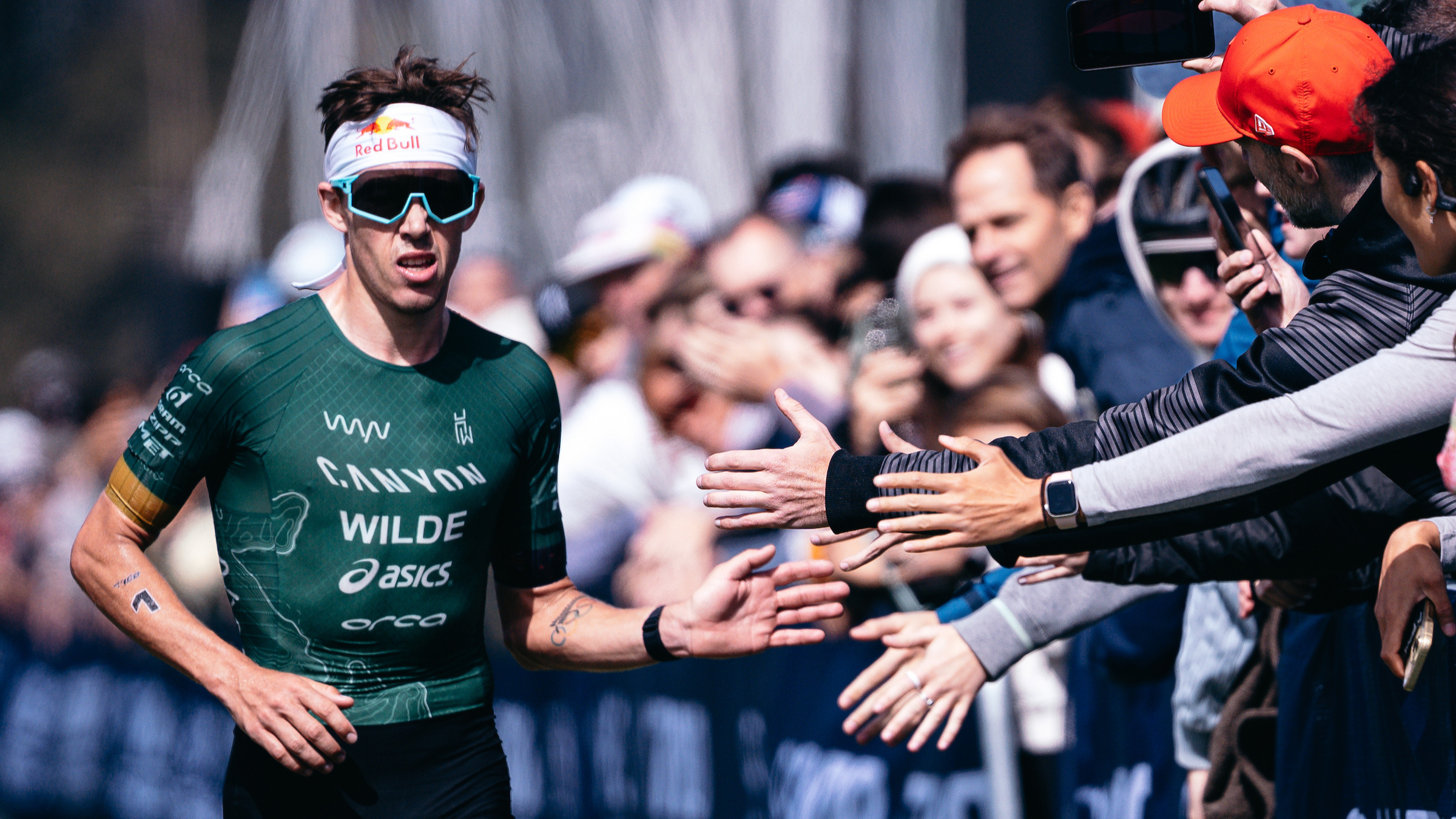Professional triathlete Hayden Wilde, wearing a dark green triathlon suit and sunglasses, greets fans with high-fives in the final chute of the race