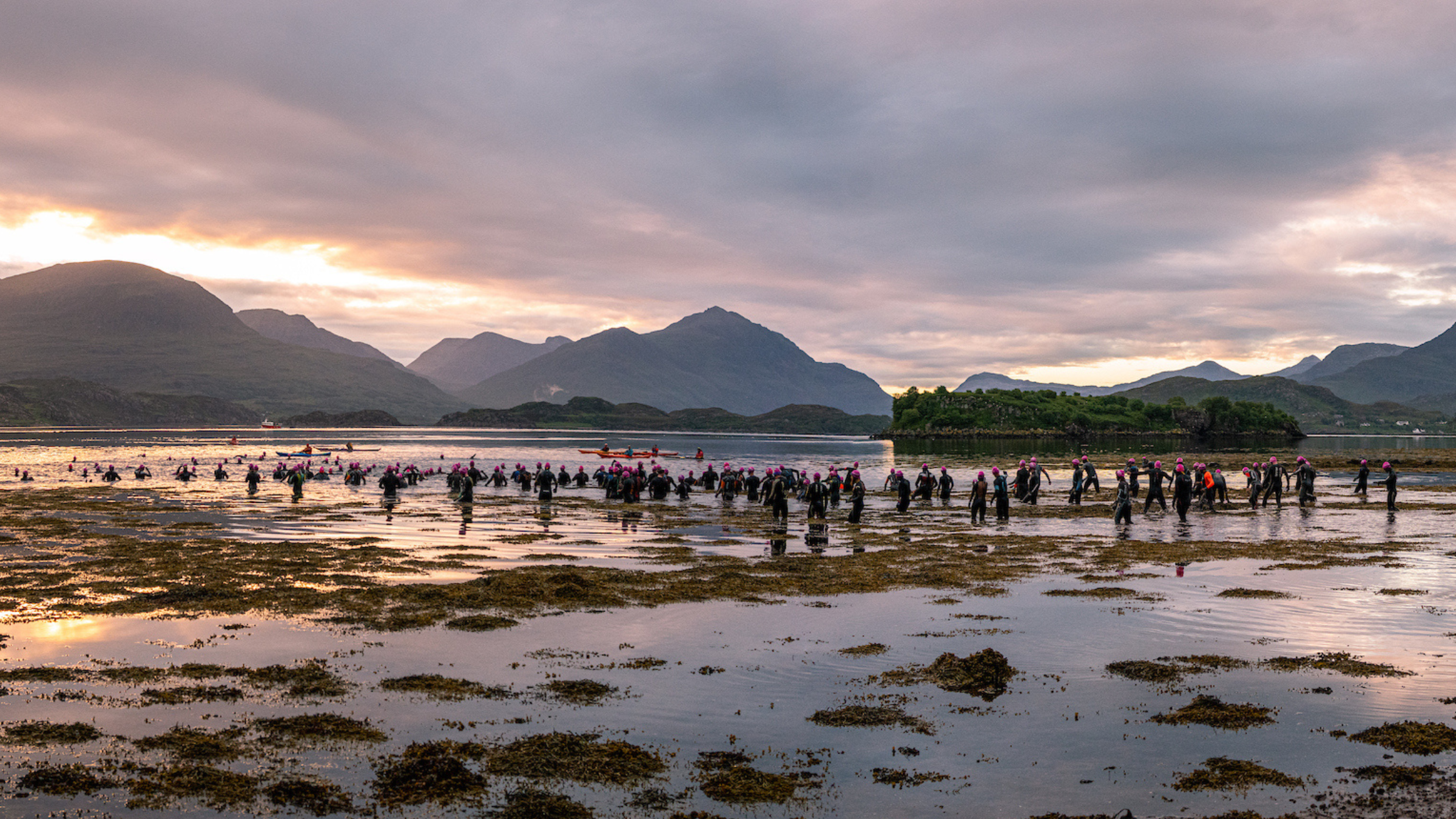 Triathletes in black wetsuits wade into the water with mountains and sunrise in the background