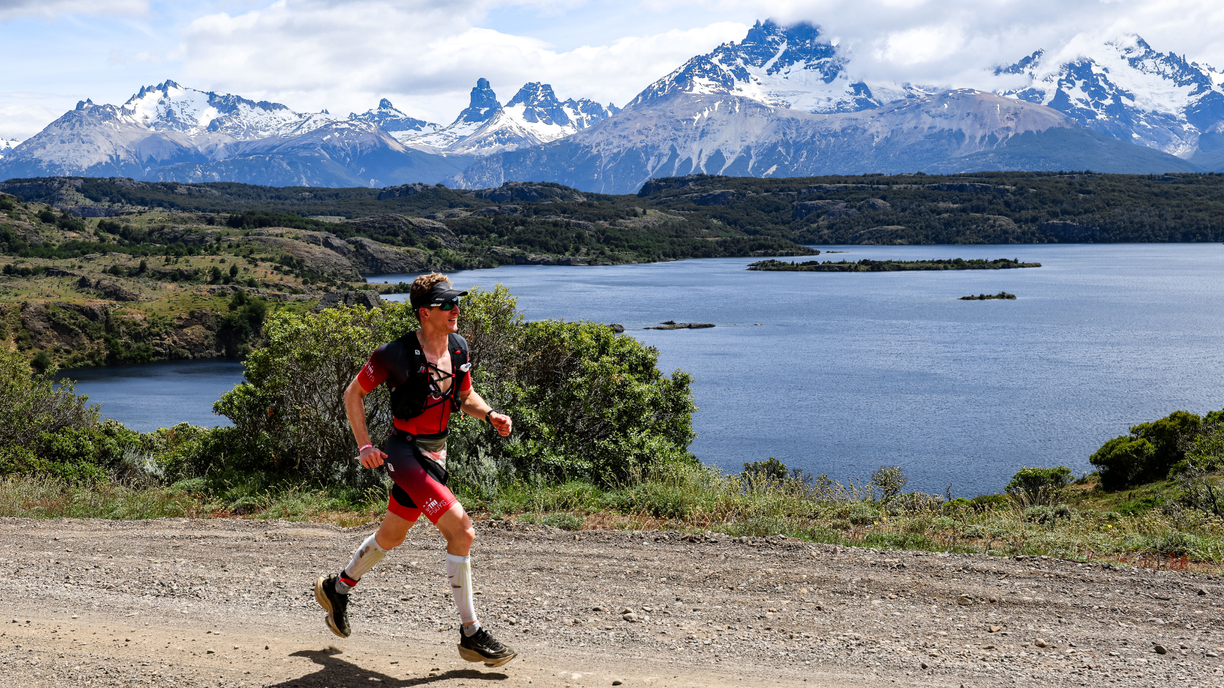 Man running in a tri suit on a path in front of a lake with snow capped mountains in the background
