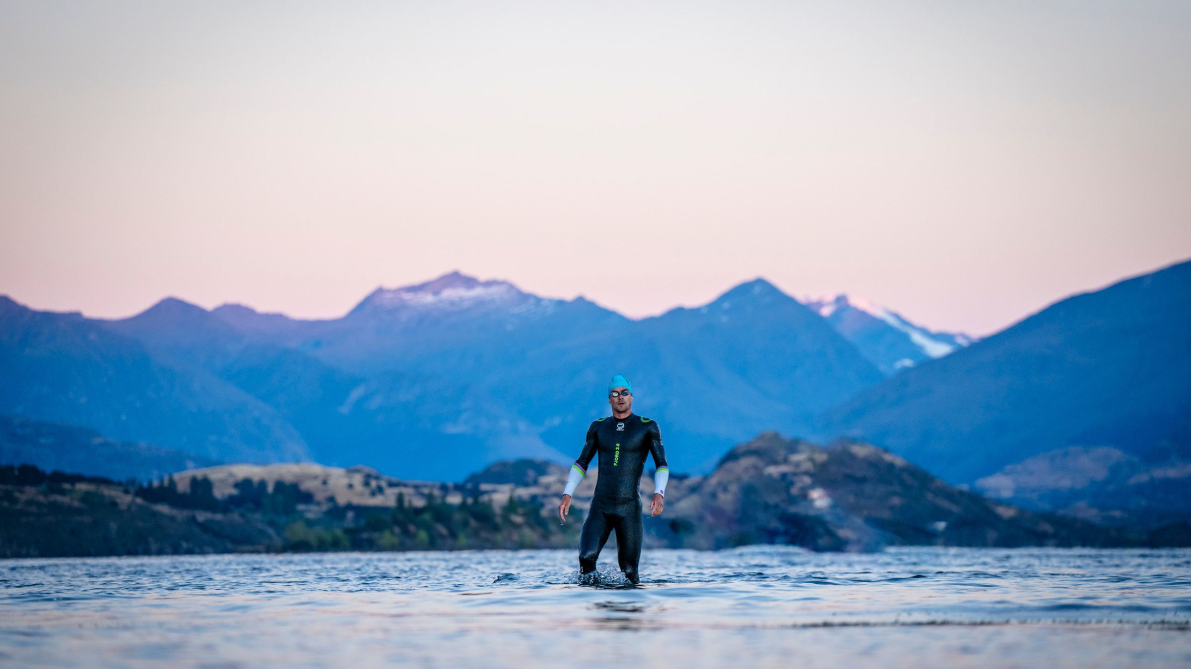 man in wetsuit wades in lake with mountains in the background