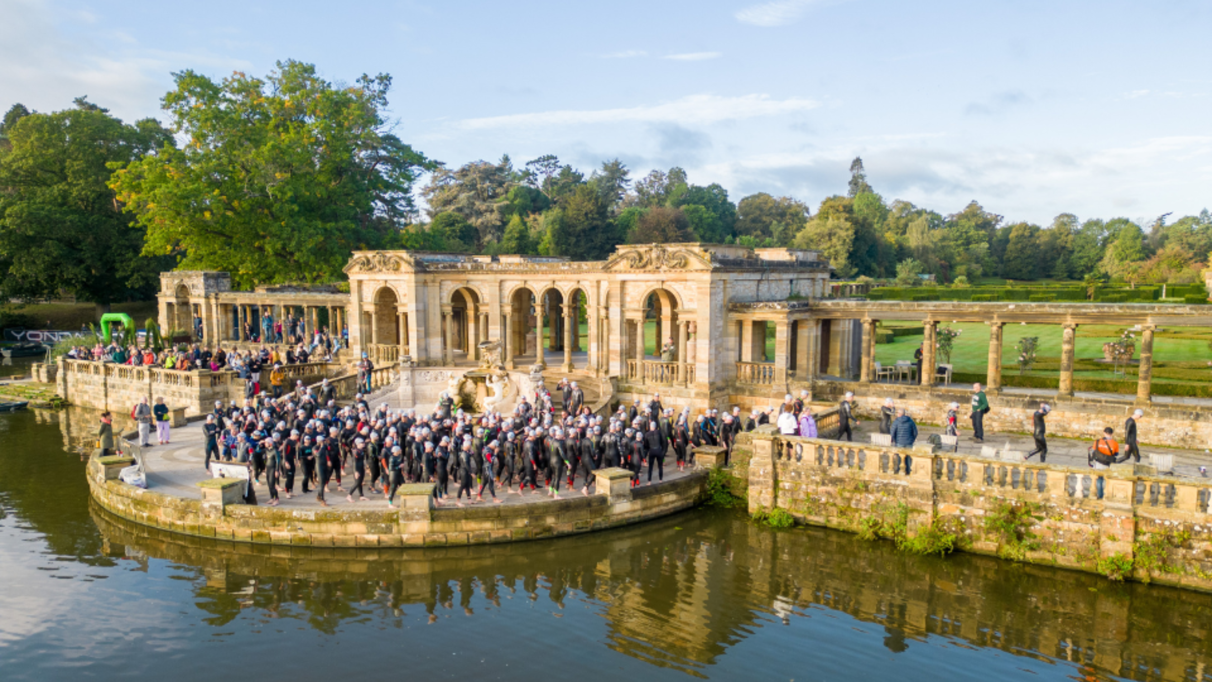 triathletes in wetsuits and swim caps line up outside of arched brick structure on castle grounds near small body of water