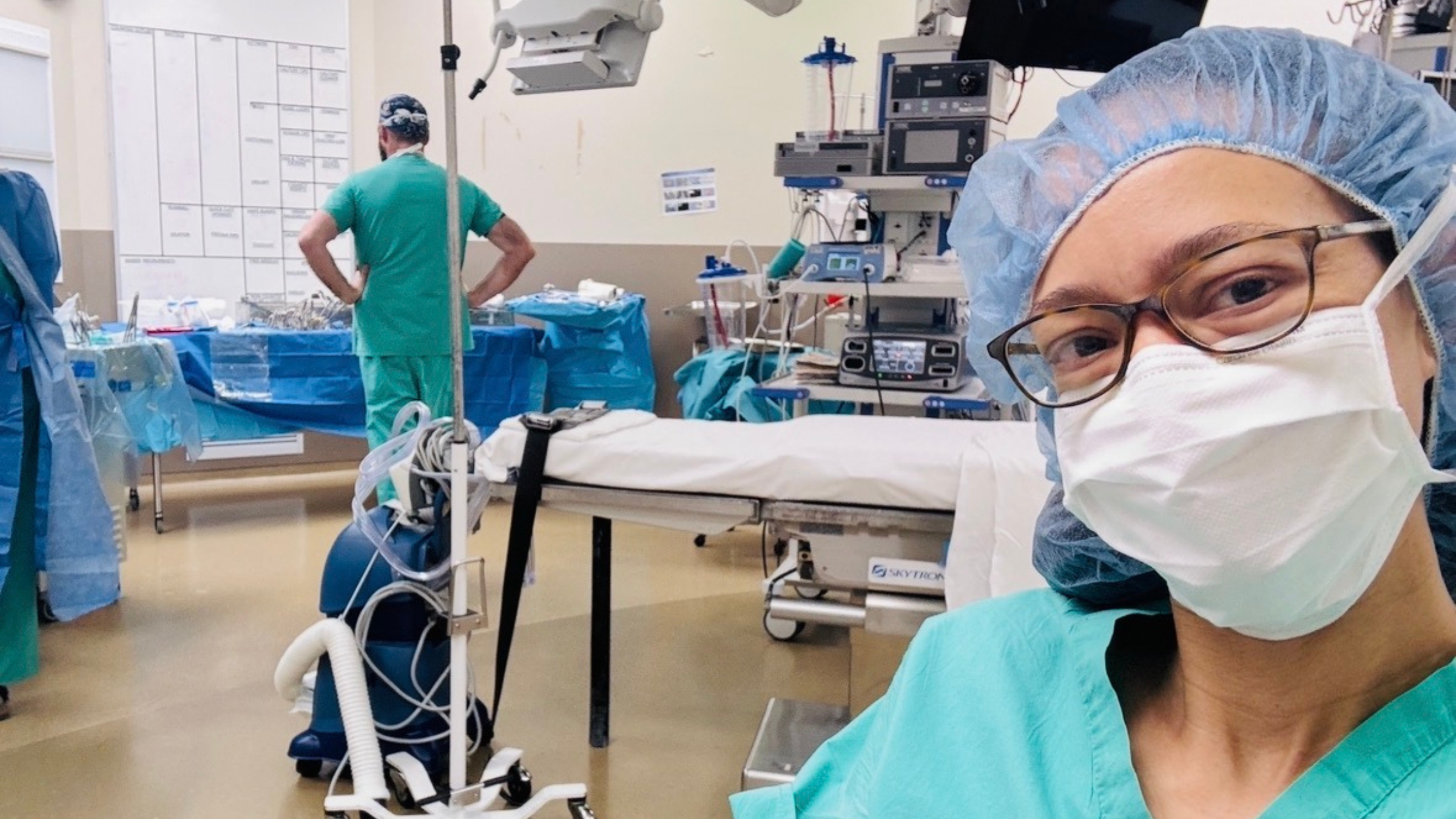 Woman dressed as a surgeon, wearing a mask and scrubs, takes a selfie in the operating room